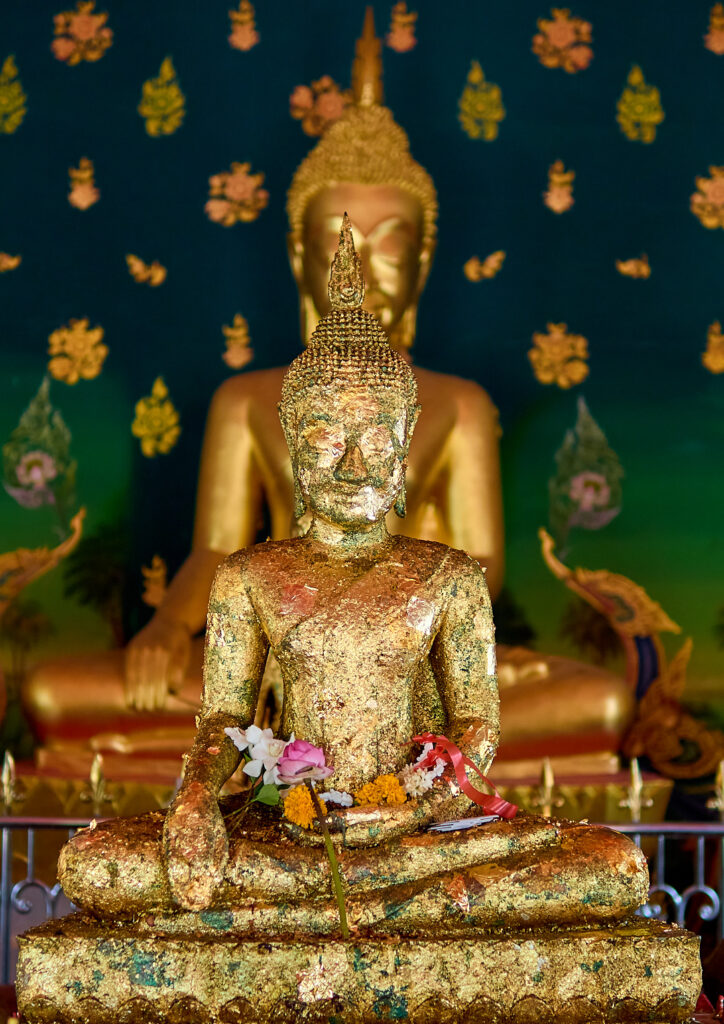 Une statue de Bouddha assise, richement recouverte de feuilles d'or et ornée de guirlandes de fleurs, est au premier plan. Derrière elle, une statue de Bouddha plus grande et dorée, légèrement floue, est visible sur un fond mural orné de motifs verts et dorés.