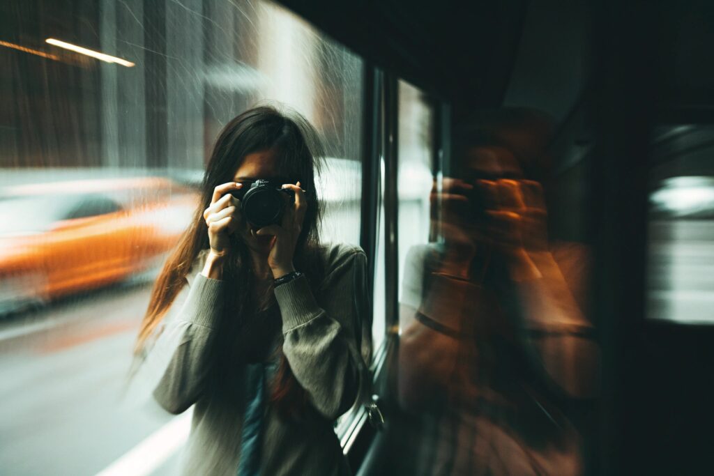 Gros plan sur une femme aux longs cheveux foncés tenant un appareil photo à hauteur des yeux, son visage partiellement caché. Elle est vue à travers une vitre mouillée ou embuée, et l'arrière-plan flou et rapide montre des lumières et la silhouette d'une voiture orange en mouvement, suggérant un déplacement rapide (train, voiture).