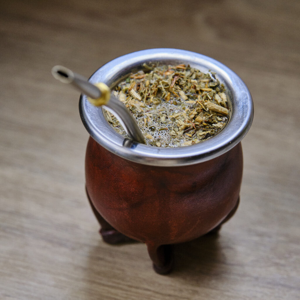 Une calebasse de maté en cuir marron et à trois pieds, avec un rebord en métal, contient du yerba maté infusé et une bombilla argentée. La boisson chaude crée de petites bulles à la surface, le tout posé sur une surface en bois clair.