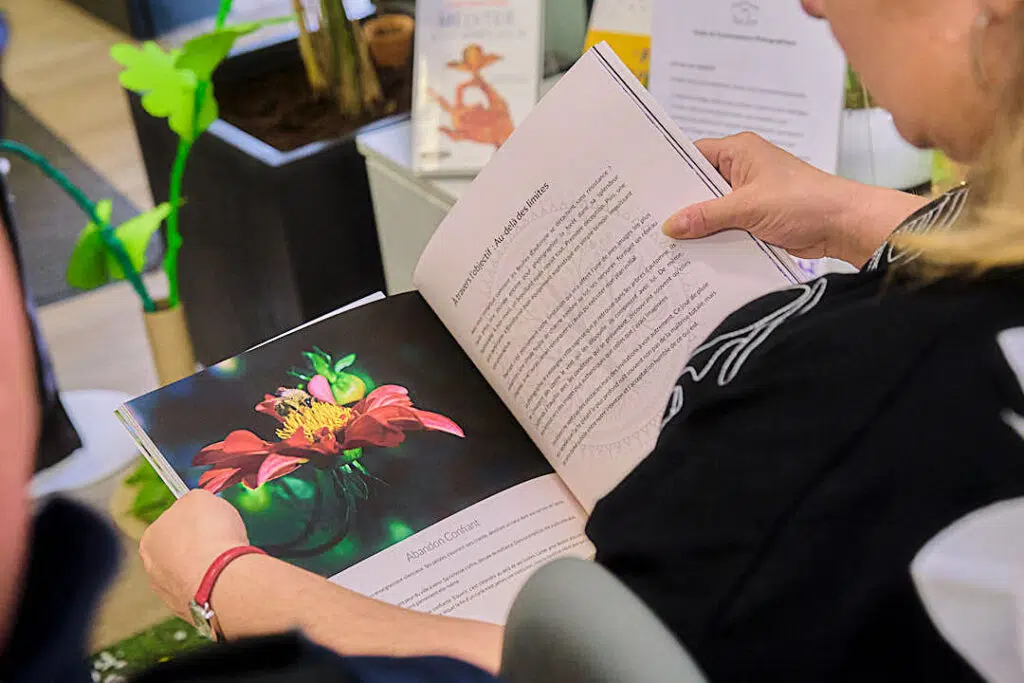 Gros plan sur une personne tenant un livre ouvert, les pages tournées vers le spectateur. La page de gauche montre une photographie d'une fleur rouge aux étamines jaunes avec une abeille dessus. La page de droite contient du texte en français. Idéal pour les thèmes de lecture, de photographie et de nature.